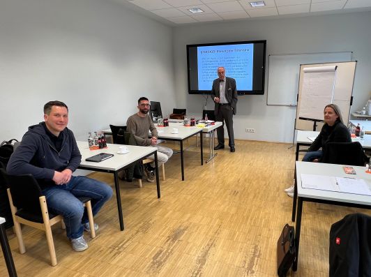 Schulungsraum mit Redner vor Bildschirm und drei Teilnehmern an Tischen