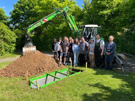 Lehrgangsteilnehmer vor Bagger an ausgehobenem Grab